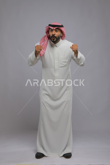 ايماءات الربح والانتصار ،حركات تدل على الفوز وانجاز المهام والكسب ،بورتريه لشاب عربي خليجي سعودي يرتدي الشماغ والزي التقليدي يرفع يديه بتفاعل وحماس ،الاندماج بتشجيع فريق كرة القدم، صورة الجسم بالطول الكامل، خلفية رمادية