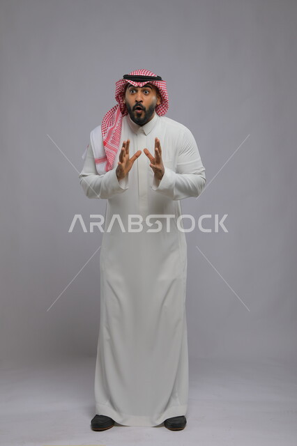 النظر إلى الكاميرا بإيماءات الذعر والخوف ,الشعور بالصدمة و الدهشة والتعجب والذهول , بورتريه لشاب عربي خليجي سعودي يرتدي الشماغ والزي التقليدي و يرفع يديه للأعلى بتعابير التفاجؤ عند رؤية شيء ما, صورة الجسم بالكامل, خلفية رمادية
