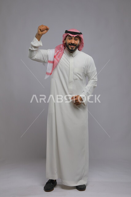 حركات تدل على القوة والنجاح، الكسب والربح وانجاز المهام، بورتريه لشاب عربي خليجي سعودي يرتدي الشماغ والثوب التقليدي و يرفع يديه للأعلى بتعابير  تحقيق الأهداف ,النظر إلى الكاميرا بإيماءات الفرح والانتصار, صورة الجسم بالطول الكامل, خلفية رمادية
