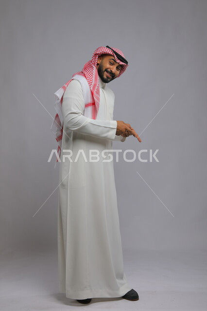  تحديد موقع معين بإيماءات السرور والبشاشة ،حركات تدل على هذا المكان (هنا)، بورتريه لشاب عربي خليجي سعودي مبتسم يرتدي الشماغ والثوب التقليدي يشير بأصابعه نحو الأسفل وينظر إلى الكاميرا، صورة الجسم بالطول الكامل، خلفية رمادية