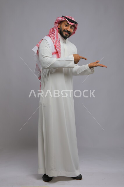 النظر إلى الكاميرا بإيماءات السعادة والسرور, بورتريه لرجل عربي خليجي سعودي مبتسم يرتدي الثوب التقليدي والشماغ يفتح كفه للأعلى ويشير إلى عنصر ما, صورة الجسم بالطول الكامل , خلفية رمادية
