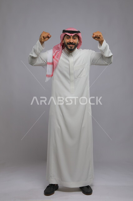 الشغف في متابعه فريق كرة القدم، حركات تدل على الكسب والربح وانجاز المهام ،ايماءات الفوز والانتصار ،بورتريه لشاب عربي خليجي سعودي يرتدي الشماغ والزي التقليدي يرفع يديه بتفاعل وحماس واندماج ،صورة الجسم بالطول الكامل، خلفية رمادية