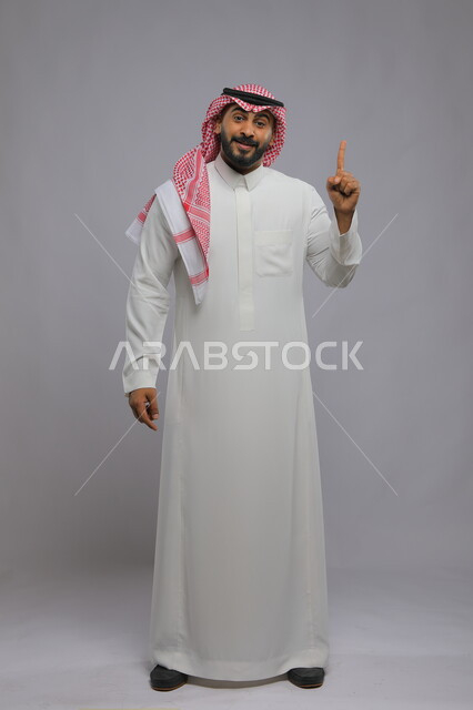 الوقوف باستقامة والنظر إلى الكاميرا، ايماءات الاستفسار والاشارة إلى شيء ما,  بورتريه لرجل عربي خليجي سعودي مبتسم يرتدي الشماغ والثوب التقليدي يرفع اصبعه للأعلى بإيماءات التوضيح, صورة الجسم بالطول الكامل , خلفية رمادية
