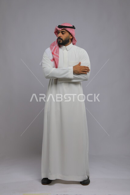 تعابير الانتظار و النظر الى شيء ما ، ايماءات التفكير ومحاولة تذكر امر ما، بورتريه لرجل عربي خليجي سعودي يرتدي الشماغ والثوب التقليدي يقف بحركات أيدي متقاطعه ويقوم بالانتظار ، صورة الجسم بالطول الكامل، خلفية رمادية 