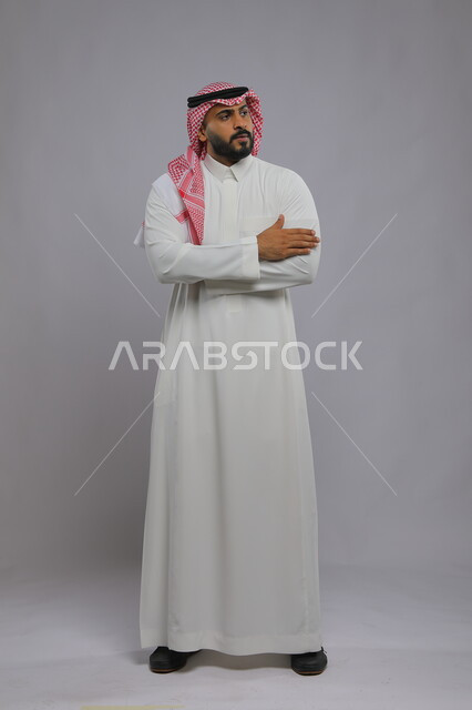 ايماءات التفكير ومحاولة تذكر امر ما، تعابير الانتظار و النظر الى شيء ما ، بورتريه لرجل عربي خليجي سعودي يرتدي الشماغ والثوب التقليدي يقف بحركات أيدي متقاطعه ويقوم بالانتظار ، صورة الجسم بالطول الكامل، خلفية رمادية 