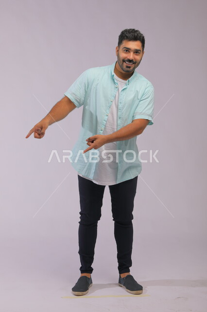  تحديد موقع معين بإيماءات السرور والبشاشة ،حركات تدل على هذا المكان (هنا)، بورتريه لشاب عربي خليجي سعودي مبتسم يرتدي الزي الكاجوال يشير بأصابعه نحو الأسفل وينظر إلى الكاميرا، صورة الجسم بالطول الكامل، خلفية رمادية.