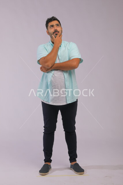 التأمل والتخطيط للمستقبل، إيجاد حل لمشكلة ما، التركيز وشرود الذهن والتمعن، بورتريه لشاب  عربي خليجي سعودي يرتدي الزي الكاجوال يضع يده على ذقنه و ينظر إلى الأعلى بإيماءات التفكير، صورة الجسم بالطول الكامل، خلفية رمادية.