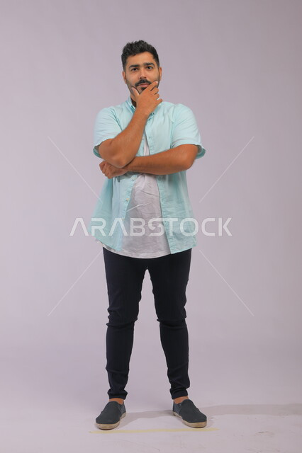 Finding a solution to a problem, contemplating and planning for the future, concentration, absent-mindedness, and contemplation, portrait of a Saudi Gulf Arab young man wearing casual clothes, placing his hand on his chin and looking at the camera with thinking gestures, full-length body portrait, gray background.