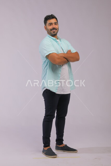 شخصية مهمة سعودية يقف متكتف اليدين، مفهوم الأناقة والرجولة والاهتمام بالمظهر الخارجي، بورتريه لرجل عربي خليجي سعودي مبتسم يرتدي اللباس الكاجوال ينظر إلى الكاميرا بإيماءات الثقه بالنفس، صورة لكامل الجسم، خلفية رمادية.