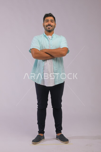 شخصية مهمة سعودية يقف متكتف اليدين، مفهوم الأناقة والرجولة والاهتمام بالمظهر الخارجي، بورتريه لرجل عربي خليجي سعودي مبتسم يرتدي اللباس الكاجوال ينظر إلى الكاميرا بإيماءات الثقه بالنفس، صورة لكامل الجسم، خلفية رمادية.