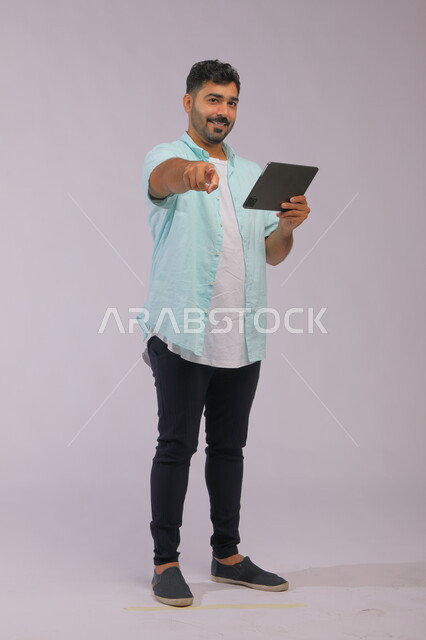 ايماءات تدل على اختيار شخص ما، استخدام التقنيه والتكنولوجيا الحديثه، بورتريه لشاب عربي خليجي سعودي مبتسم يرتدي الزي الكاجوال يشير بأصبع السبابة للأمام و ينظر إلى الكاميرا، تصفح مواقع التواصل الاجتماعي عبر  جهاز التابليت، صورة الجسم بالكامل، خلفية رمادية