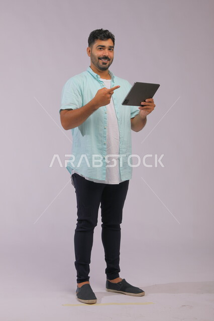 ايماءات الاعجاب باستخدام التقنيه والتكنولوجيا الحديثه، بورتريه لشاب عربي خليجي سعودي مبتسم يرتدي الزي الكاجوال يشير بأصبع السبابة على جهاز التابليت و ينظر إلى الكاميرا، تصفح مواقع التواصل الاجتماعي عبر الأجهزة اللوحية، صورة الجسم بالكامل، خلفية رمادية