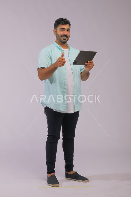 Gestures of acceptance and satisfaction, integrating technology and technology into practical life, portrait of a smiling Saudi Gulf Arab young man wearing casual clothes, raising his thumb up with the quality sign and looking at the camera, browsing social media sites via a tablet, full body photo, gray background