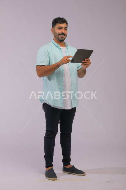 دمج التقنية والتكنولوجيا في الحياة العملية، عقد اجتماع عمل عن بعد ،الدردشة مع الاصدقاء عبر الانترنت، بورتريه لشاب عربي خليجي سعودي مبتسم يرتدي الزي الكاجوال ينظر إلى الكاميرا، متابعة اخر أخبار السوشال ميديا باستخدام جهاز التابليت، صورة الجسم بالكامل، خلفية رمادية
