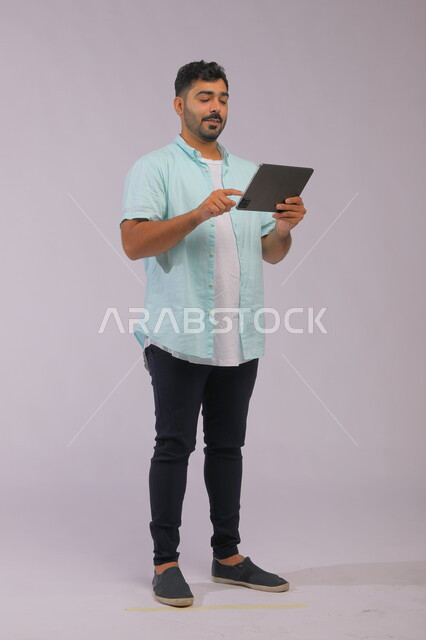  إستخدام برامج الاجهزة الذكية، ايماءات الاندماج والتركيز ،الدردشة والتحدث مع الاصدقاء عبر الانترنت، بورتريه لشاب عربي خليجي سعودي مبتسم يرتدي الزي الكاجوال يقوم بمتابعة اخبار وسائل التواصل الاجتماعي باستخدام جهاز التابليت، صورة الجسم بالكامل، خلفية رمادية