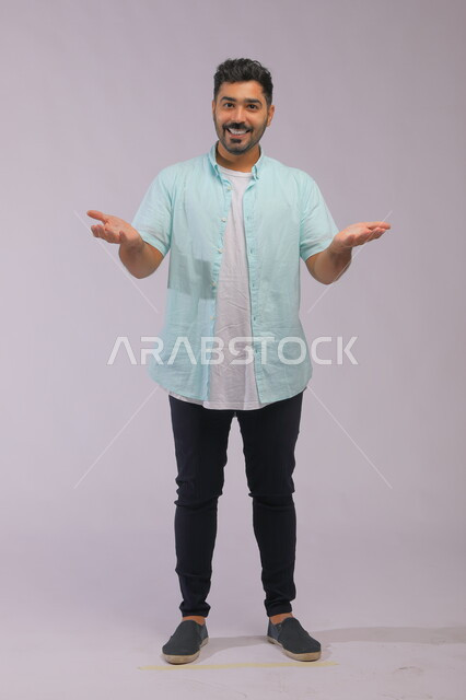 الإعجاب والمدح بشيء جميل, الشكر والامتنان على المعروف، التساؤل عن حدوث أمر ما، بورتريه لشاب عربي سعودي خليجي يرتدي الزي الكاجوال يرفع كفيه وينظر إلى الكاميرا، انتظار شيء ما، صورة الجسم بالطول الكامل, خلفية رمادية.