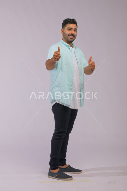 الشعور بالإعجاب والرضا والسعاده، النظر إلى الكاميرا بايماءات القبول والموافقه, بورتريه لشاب وسيم عربي خليجي سعودي مبتسم يرتدي الزي الكاجوال ويرفع إصبع الابهام بعلامة الجودة، صورة الجسم بالطول الكامل، خلفية رمادية.