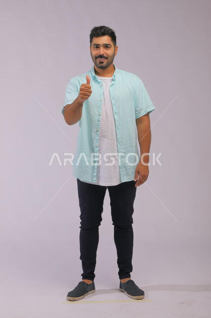 الشعور بالإعجاب والرضا والسعاده، النظر إلى الكاميرا بايماءات القبول والموافقه, بورتريه لشاب وسيم عربي خليجي سعودي مبتسم يرتدي الزي الكاجوال ويرفع إصبع الابهام بعلامة الجودة، صورة الجسم بالطول الكامل، خلفية رمادية.