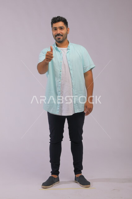 الشعور بالإعجاب والرضا والسعاده، النظر إلى الكاميرا بايماءات القبول والموافقه, بورتريه لشاب وسيم عربي خليجي سعودي مبتسم يرتدي الزي الكاجوال ويرفع إصبع الابهام بعلامة الجودة، صورة الجسم بالطول الكامل، خلفية رمادية.