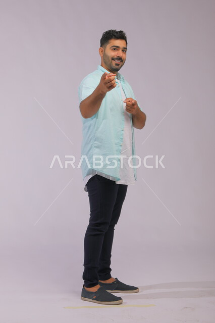 ايماءات التأكيد وتحديد الشيء المقابل, الإشارة الى شخص ما, بورتريه لشاب عربي خليجي سعودي وسيم يرتدي الي الكاجوال يرفع اصبعه السبابة للأمام بإيماءات وجه تدل على السرور, صورة الجسم بالكامل, خلفية رمادية.