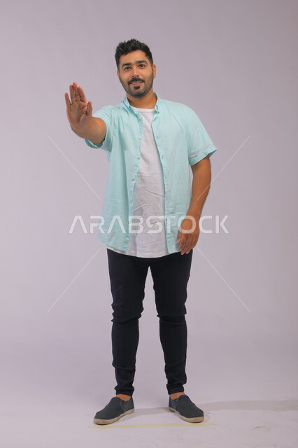 إيماءات طلب التوقف والابتعاد، الامتناع عن فعل شيء ما، الوقوف باستقامة والنظر إلى الكاميرا, بورتريه لرجل عربي خليجي سعودي يرتدي اللباس الكاجوال يرفع يده للأمام, صورة الجسم بالطول الكامل, خلفية رمادية.