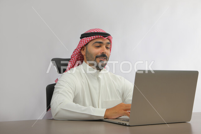 عقد اجتماع عمل عن بعد، استخدام التقنيات الحديثة المتطورة, بورتريه لرجل عربي خليجي سعودي يرتدي الزي التقليدي يجلس أمام جهاز الحاسوب، الدردشة مع الأهل والأصدقاء, متابعه آخر أخبار السوشال ميديا، تطبيقات وبرامج الأجهزة اللوحية، خلفية رمادية.