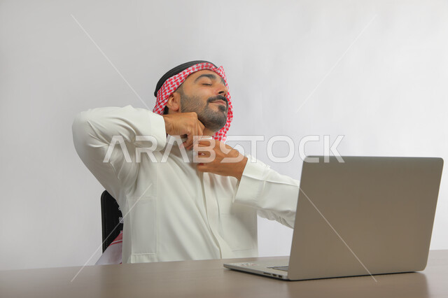 استخدام التقنيات الحديثة المتطورة, متابعه آخر أخبار السوشال ميديا، عقد اجتماع عمل عن بعد، بورتريه لرجل عربي خليجي سعودي يرتدي الزي التقليدي يجلس على المكتب أمام اللابتوب بإيماءات تدل على التعب والنعاس، الدردشة مع الأهل والأصدقاء, خلفية رمادية.