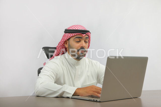 استخدام التقنيات الحديثة المتطورة, متابعه آخر أخبار السوشال ميديا، عقد اجتماع عمل عن بعد، بورتريه لرجل عربي خليجي سعودي يرتدي الزي التقليدي يجلس على المكتب أمام اللابتوب بإيماءات تدل على التعب والنعاس، الدردشة مع الأهل والأصدقاء, خلفية رمادية.