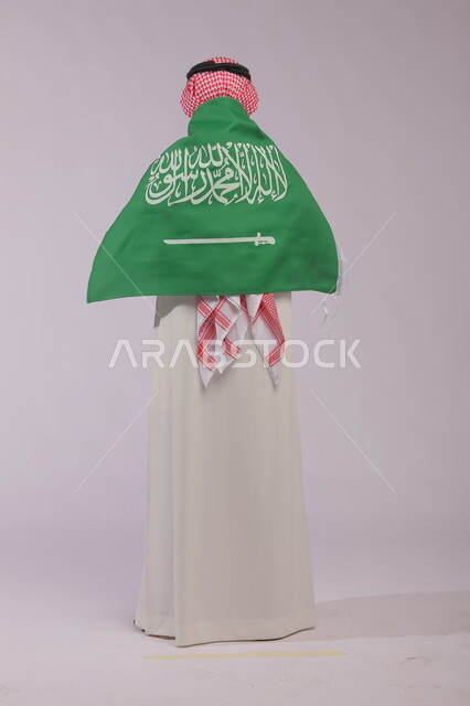 مناسبات شعبيه وطنية، اظهار الحب والولاء للملكه، احياء ذكرى اليوم الوطني السعودي 23 سبتمبر، بورتريه من الخلف لرجل عربي خليجي سعودي يرتدي الشماغ والثوب التقليدي  ويضع علم السعودية على كتفيه في العيد الوطني، يوم العلم 11 مارس ، صورة الجسم بالكامل ،خلفية رمادية