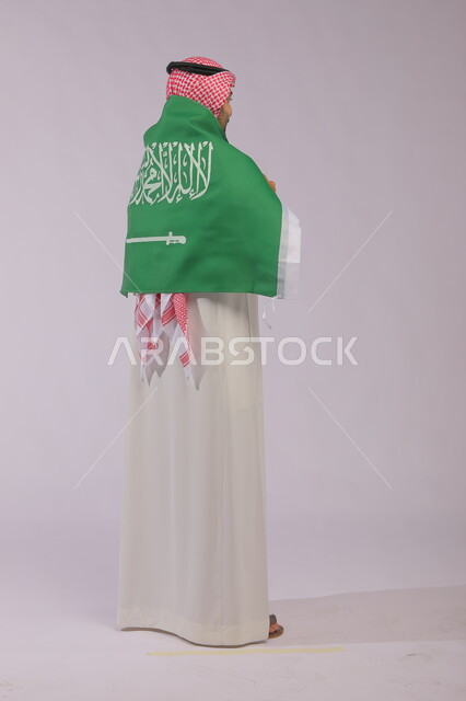 Popular national occasions, showing love and loyalty to the Queen, commemorating the Saudi National Day, September 23, a portrait from the back of a Saudi Gulf Arab man wearing a shemagh and traditional thobe and placing the Saudi flag on his shoulders on National Day, Flag Day, March 11, full body portrait, gray background