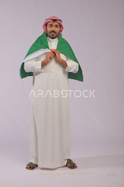 الفخر والاعتزاز بالهوية الوطنية، الوقوف باستقامة ووضع علم المملكة على كتفيه بإيماءات السرور، اليوم الوطني السعودي 23 سبتمبر، بورتريه لشاب عربي خليجي سعودي مبتسم يرتدي الشماغ والثوب التقليدي وينظر إلى الكاميرا ،يوم العلم 11 مارس، صورة الجسم بالكامل، خلفية رمادية