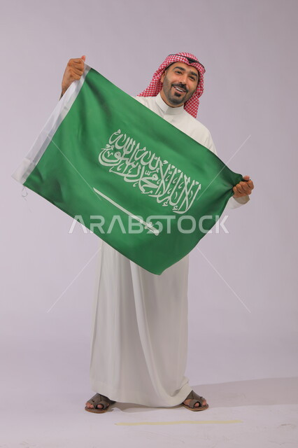 رفع علم المملكة بإيماءات البهجة و الضحك، إظهار الفخر والولاء للبلاد وتعزيز الروح الوطنية، اليوم الوطني السعودي 23 سبتمبر، بورتريه لشاب عربي خليجي سعودي مبتسم يرتدي الشماغ والثوب التقليدي وينظر إلى الكاميرا ،يوم العلم 11 مارس، صورة الجسم بالكامل، خلفية رمادية