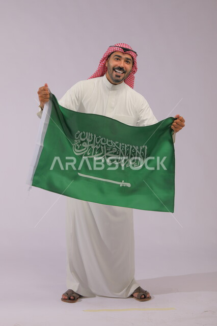 Remembering history and enhancing the patriotic spirit, displaying the Kingdom’s flag with gestures of laughter and joy, Saudi National Day, September 23, portrait of a smiling Gulf Arab Saudi man wearing a shemagh and traditional thobe and looking at the camera, Flag Day, March 11, full body portrait, gray background