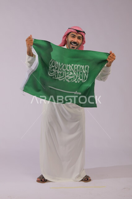 عرض علم المملكة بإيماءات الضحك والبهجة، إظهار الفخر والولاء للبلاد وتعزيز الروح الوطنية، اليوم الوطني السعودي 23 سبتمبر، بورتريه لشاب عربي خليجي سعودي مبتسم يرتدي الشماغ والثوب التقليدي وينظر إلى الكاميرا ،يوم العلم 11 مارس، صورة الجسم بالكامل، خلفية رمادية