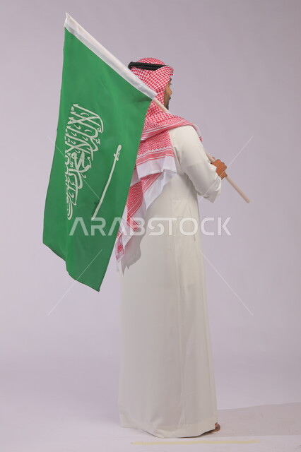 الوقوف باعتزاز وفخر بالهوية الوطنية، اليوم الوطني السعودي 23 سبتمبر، بورتريه من الخلف لشاب عربي خليجي سعودي يرتدي الثوب والشماغ التقليدي يحمل بيده علم المملكة العربيه السعودية وينظر إلى شيء ما، يوم العلم 11 مارس، صورة الجسم بالطول الكامل، خلفية رمادية