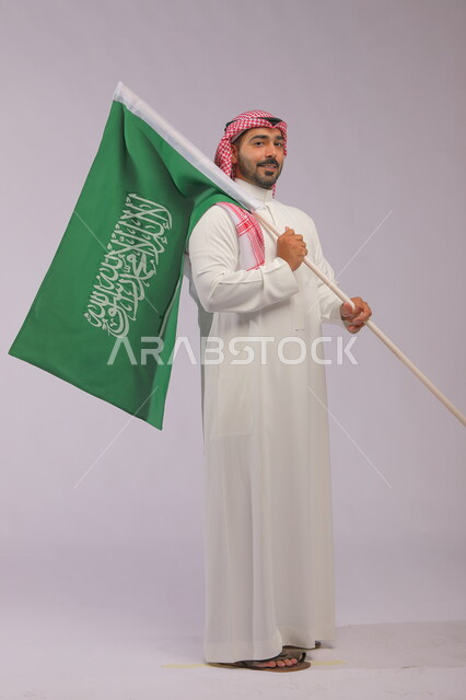Pride and pride in national identity, Saudi National Day, September 23, portrait of a smiling Saudi Gulf Arab young man wearing the traditional thobe and shemagh, holding the flag of the Kingdom of Saudi Arabia in his hand and looking at the camera, Flag Day, March 11, full-length body portrait, gray background