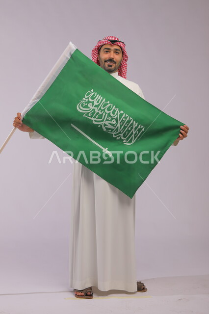 عرض علم المملكة بإيماءات الحب و السرور، إظهار الفخر والولاء للبلاد وتعزيز الروح الوطنية، اليوم الوطني السعودي 23 سبتمبر، بورتريه لشاب عربي خليجي سعودي مبتسم يرتدي الشماغ والثوب التقليدي وينظر إلى الكاميرا ،يوم العلم 11 مارس، صورة الجسم بالكامل، خلفية رمادية