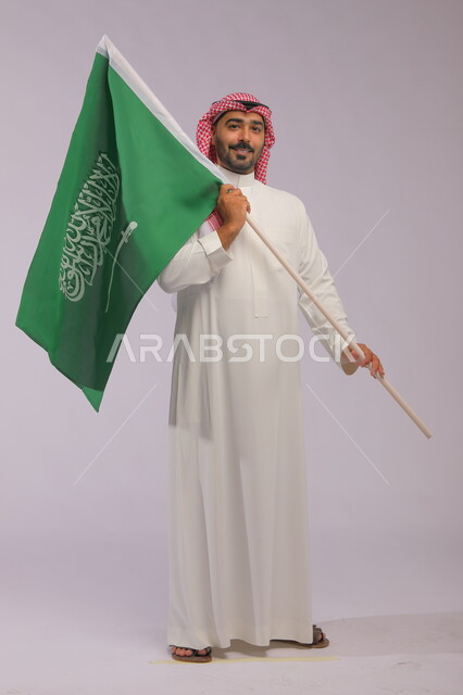 يوم العلم 11 مارس، الاحتفال باليوم الوطني السعودي 23 سبتمبر والعيد الوطني، بورتريه لرجل عربي خليجي سعودي يرتدي الشماغ والثوب التقليدي يحمل بيده علم المملكة وينظر إلى الكاميرا، الفخر والاعتزاز بامتلاك الهوية الوطنية، صورة الجسم بالطول الكامل، خلفية رمادية