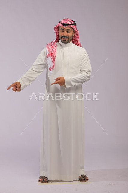  إشارات وحركات يدين تدل على هذا المكان (هنا)، تحديد موقع معين بإيماءات السرور والبشاشة، بورتريه لرجل عربي خليجي سعودي مبتسم يرتدي الزي التقليدي والشماغ يشير بأصابعه نحو الأسفل، صورة الجسم بالطول الكامل، خلفية رمادية.