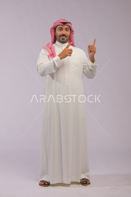الوقوف باستقامة والنظر إلى الكاميرا، إشارات وحركات يدين تدل على هذا المكان (هنا)، تحديد موقع معين بإيماءات السرور والبشاشة، بورتريه لرجل عربي خليجي سعودي مبتسم يرتدي الزي التقليدي والشماغ يشير بأصابعه نحو الأعلى، صورة الجسم بالطول الكامل، خلفية رمادية.