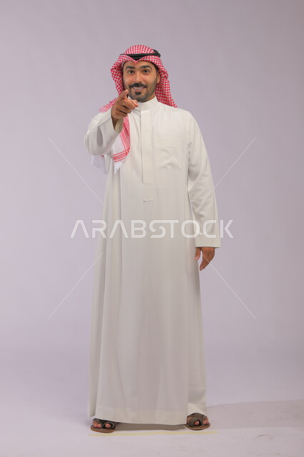 حركات وإيماءات جسد تدل على الإشارة لشخص ما، الوقوف باستقامة والنظر إلى الكاميرا, بورتريه لرجل عربي خليجي سعودي يرتدي الزي التقليدي يشير بإصبعه للأمام, صورة الجسم بالطول الكامل, خلفية رمادية.
