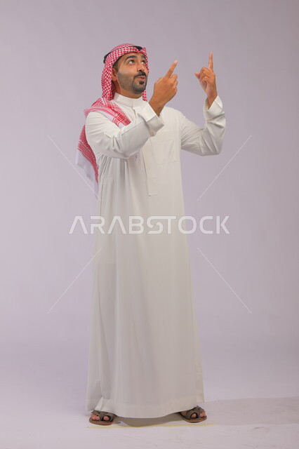 Identifying a specific location with gestures indicating astonishment and wonder, signs and hand movements indicating this place (here), a portrait of a Saudi Gulf Arab man wearing a traditional dress and a shemagh, pointing with his fingers upward, a full-length body image, a gray background.