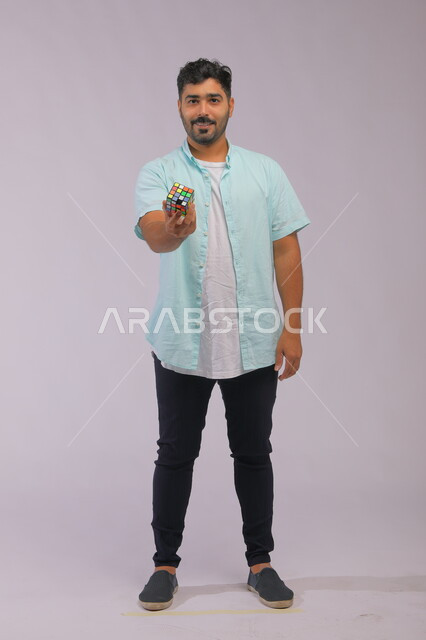 عرض تجربة اللعب بالروبيك كيوب، نشاطات تقوية الذاكره ،بورتريه لشاب وسيم عربي خليجي سعودي يرتدي الزي الكاجوال ويمسك بيده مكعب مغناطيس السرعه ،الوقوف باستقامة والنظر إلى الكاميرا، قضاء أوقات ممتعه في العطلة الصيفية، صوة الجسم بالطول الكامل، خلفية رمادية