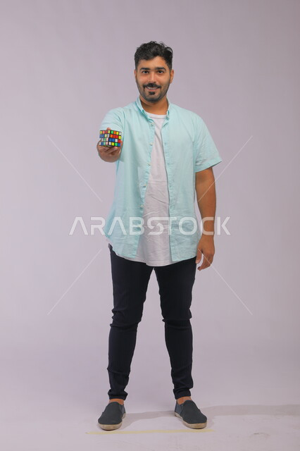 ألعاب تعليمية ترفيهيه، نشاطات تقوية الذاكره ،بورتريه لشاب وسيم عربي خليجي سعودي يرتدي الزي الكاجوال ويمسك بيده مكعب مغناطيس السرعه ،الوقوف باستقامة وتقديم روبيك كيوب للتجربة، قضاء أوقات ممتعه في العطلة الصيفية، صوة الجسم بالطول الكامل، خلفية رمادية