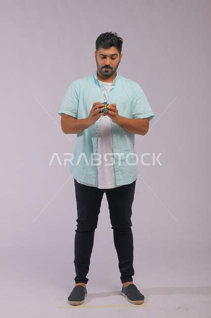 ألعاب تنمية الذكاء والمهارات العقليه، نشاطات تقوية الذاكره ،بورتريه لشاب وسيم عربي خليجي سعودي يرتدي الزي الكاجوال ويمسك بيده المكعب السحري ،الوقوف باستقامة بإيماءات التركيز والاندماج، قضاء أوقات ممتعه في العطلة الصيفية، صوة الجسم بالطول الكامل، خلفية رمادية