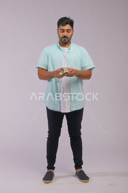 نشاطات تقوية الذاكره ،بورتريه لشاب وسيم عربي خليجي سعودي يرتدي الزي الكاجوال ويمسك بيده المكعب السحري ،ألعاب تنمية الذكاء والمهارات  العقليه، الوقوف باستقامة بإيماءات التركيز والاندماج، قضاء أوقات ممتعه في العطلة الصيفية، صوة الجسم بالطول الكامل، خلفية رمادية
