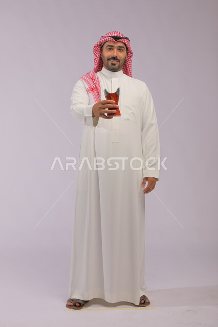 التطيب بالعود والبخور, استخدام خشب العود بالأعياد والمناسبات, بورتريه لرجل عربي خليجي سعودي مبتسم يرتدي الثوب التقليدي والشماغ ينظر إلى الكاميرا ويحمل المبخرة, الاهتمام بالمظهر الخارجي, العادات والتقاليد السعودية, صورة الجسم بالطول الكامل, خلفية رمادية