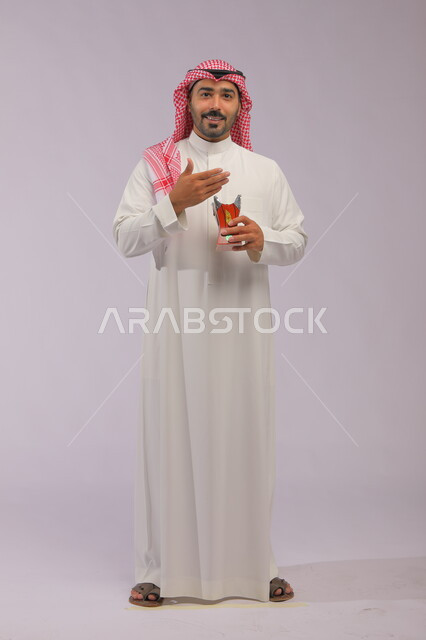 التطيب بالعود والبخور, استخدام خشب العود بالأعياد والمناسبات, بورتريه لرجل عربي خليجي سعودي مبتسم يرتدي الثوب التقليدي والشماغ ينظر إلى الكاميرا ويحمل المبخرة, الاهتمام بالمظهر الخارجي, العادات والتقاليد السعودية, صورة الجسم بالطول الكامل, خلفية رمادية