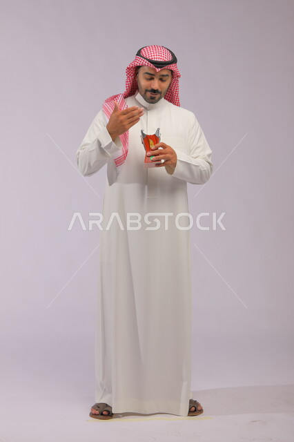 العادات والتقاليد السعودية, التطيب بالعود والبخور, استخدام خشب العود بالأعياد والمناسبات, بورتريه لرجل عربي خليجي سعودي مبتسم يرتدي الثوب التقليدي والشماغ يقوم بالتعطر من حامل البخور, الاهتمام بالمظهر الخارجي, صورة الجسم بالطول الكامل, خلفية رمادية