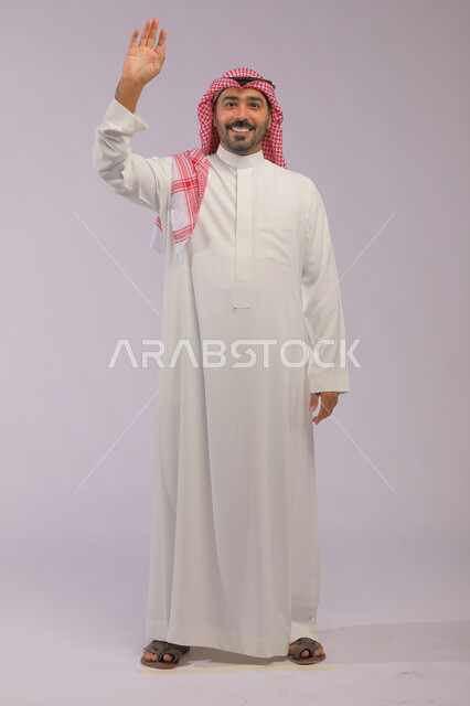 ايماءات إلقاء التحية والسلام والسعادة بالترحيب، بورتريه لرجل عربي خليجي سعودي يرتدي الثوب والشماغ التقليدي يقوم برفع يده اليمنى للأعلى، صورة الجسم بالطول الكامل، خلفية رمادية.